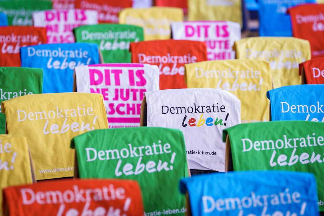 Das Bild zeigt die für die Gäste vorbereiteten Stuhlreihen der 4. Jahreskonferenz des Demokratie-Zentrums. Die mit bunten Stoffbeuteln vom »Bündnis Demokratie leben!« behangenen Stuhllehnen bilden eine vielfältiges und kunterbuntes Muster.