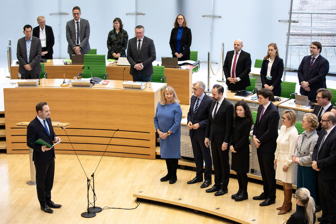 Die Ministerinnen und Minister stehen im Landtag. Der Präsident des Landtags steht am Mikrofon und spricht