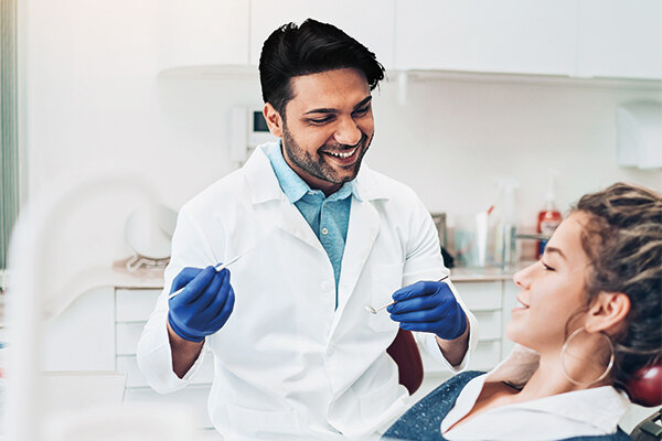 Zahnarzt blickt lächelnd auf Patienten hinunter, in der Hand hält er ein Dentalinstrument.