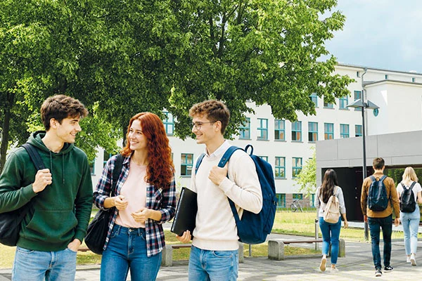 Drei junge Studierende mit Rucksäcken unterhalten sich lächelnd auf einem grünen Universitätscampus, während im Hintergrund weitere Personen vor einem Universitätsgebäude laufen.