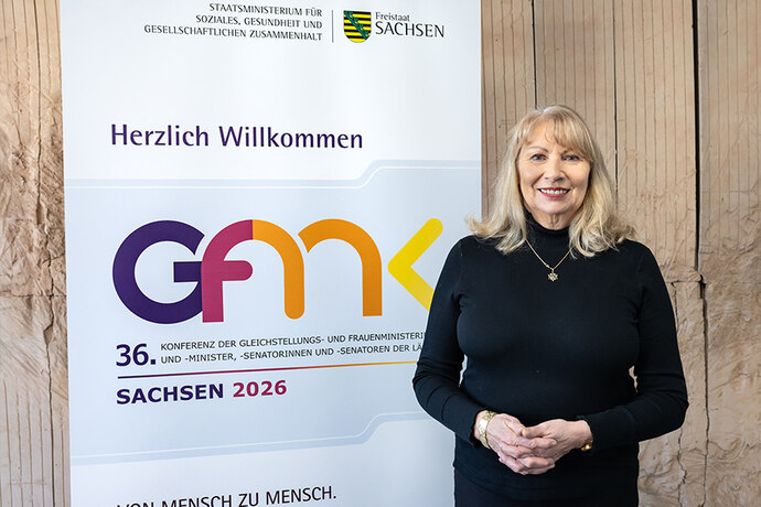 Frau steht lächelnd vor einem Roll-up mit der Aufschrift „Herzlich Willkommen“ und dem Logo „GFMK“. Darunter: „36. Konferenz der Gleichstellungs- und Frauenminister…“, „Sachsen 2026“ sowie der Slogan „Von Mensch zu Mensch.“
