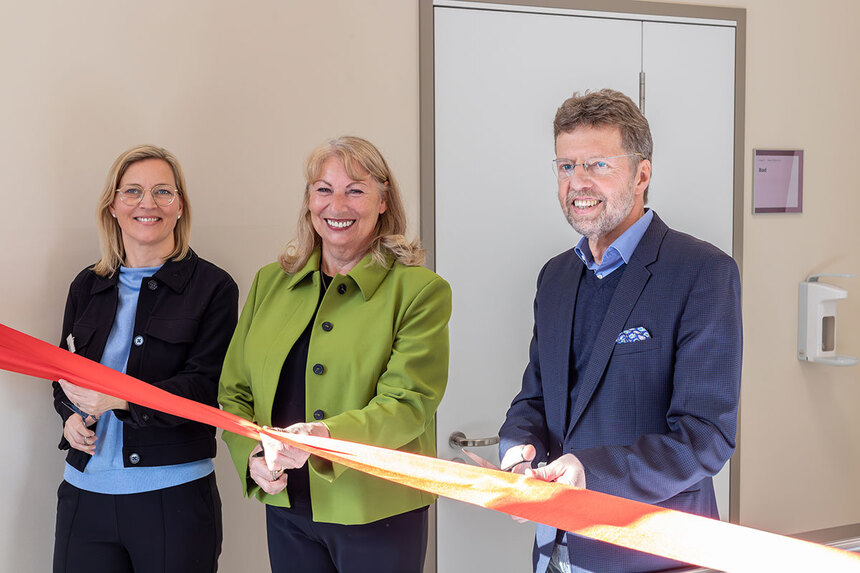 Drei Personen stehen nebeneinander vor einer Tür und schneiden gemeinsam ein rotes Band zur Einweihung eines Klinikbereichs durch.