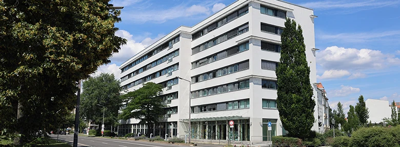 Foto des weißen Sozialministeriums. Davor eine leere Straße, Bäume und blauer Himmel.