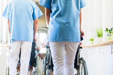 Two nurses push two patients in a wheelchair down a corridor