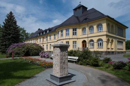 Zu sehen ist das Verwaltungsgebäude des Landeskrankenhauses Großschweidnitz, im Vordergrund eine Stele.