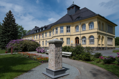 Zu sehen ist das Verwaltungsgebäude des Landeskrankenhauses Großschweidnitz, davor eine Stele.