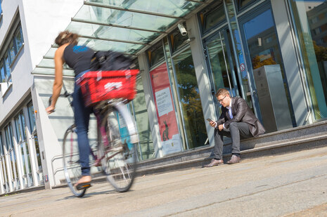 Blick auf den Haupteingang des Sozialministeriums mit vorbeifahrender Radfahrerin und einem auf den Treppenstufen sitzenden jungen Mann