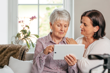 Eine jüngere Frau sitzt mit einer älteren zusammen, sie schauen gemeinsam auf einem Tablet in die Pflegedatenbank. 