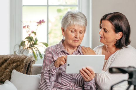 Eine ältere Frau sitze auf einer Couch und hat ein Tablet in der Hand. Neben ihr sitzt eine jüngere Frau, die den Arm um sie legt.