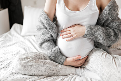 Frau sitzt auf einem Bett. Man sieht deutlichen den Schwangerschaftsbauch, den sie mit beiden Händen festhält.