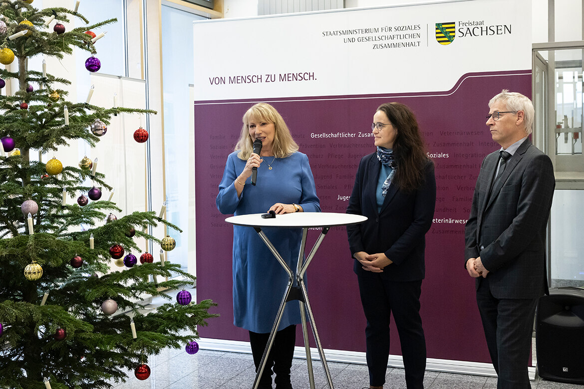 Staatsministerin Petra Köpping, Staatssekretärin Dagmar Neukirch und Rüdiger Raulfs stehen im Foyer an einem Tisch und sprechen. Daneben steht ein Weihnachtsbaum