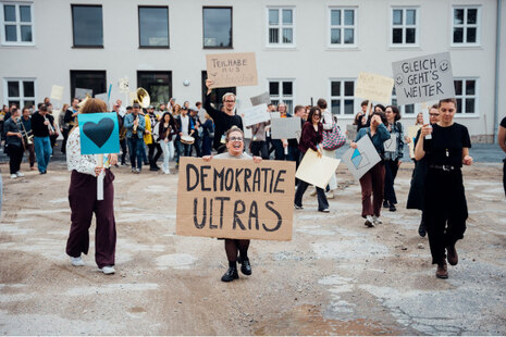 Viele junge Menschen demonstrieren vor einem großen weißen Gebäude. In der Mitte des Bildes hält eine junge Frau mit Brille ein Schild in den Händen, auf dem steht »Demokratie Ultras«.
